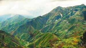 Vue panoramique des montagnes verdoyantes et escarpées de Kenscoff en Haïti, montrant des routes sinueuses à flanc de colline et des habitations dispersées sous un ciel nuageux.