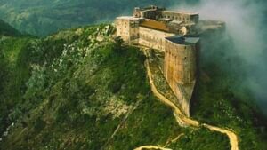 Vue aérienne de la Citadelle Laferrière, une immense forteresse en pierre grise située sur une crête montagneuse verdoyante en Haïti, partiellement entourée de brume.