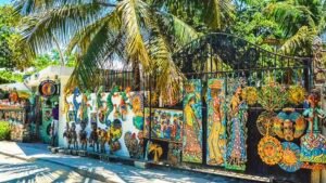 Une rue ensoleillée au Village Artistique de Noailles en Haïti, montrant des murs et des portails en fer forgé ornés de sculptures en métal découpé aux couleurs vives, sous des palmiers.