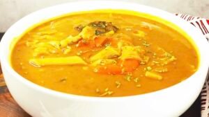 Un bol blanc rempli de soupe joumou haïtienne, une soupe de giraumon jaune orangé épaisse contenant des morceaux de viande, des carottes et des pâtes, parsemée de fines herbes.