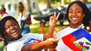 Deux jeunes femmes haïtiennes souriantes célébrant la Fête du Drapeau. L'une porte un t-shirt avec le drapeau haïtien et l'autre fait un signe de la main en tenant un petit drapeau bleu et rouge.