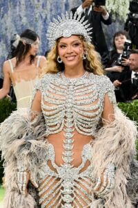 Photo de Beyoncé au Met Gala 2026. Elle porte une robe transparente ornée d'un squelette étincelant en diamants qui souligne son buste et son bassin. Elle arbore une couronne argentée majestueuse en forme de rayons et des gants squelettiques assortis. Une imposante étole en plumes gris perle entoure ses bras, sur un fond de mur floral.