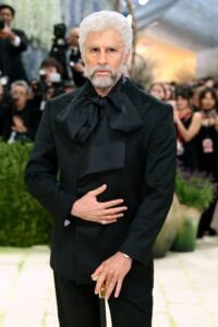 Bad Bunny méconnaissable au Met Gala 2026, vieilli grâce à des prothèses faciales ultra-réalistes. Il arbore des cheveux blancs frisés, une barbe poivre et sel et des rides marquées. Il est vêtu d'un costume noir élégant avec un nœud lavallière imposant et tient une canne à pommeau doré.