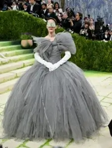 Sarah Paulson au Met Gala 2026. Elle porte une robe de bal monumentale en tulle gris anthracite avec un nœud imposant au niveau du buste et de longs gants blancs. Son visage est partiellement masqué par une paire de lunettes rectangulaires sur lesquelles sont fixés de véritables billets de un dollar.
