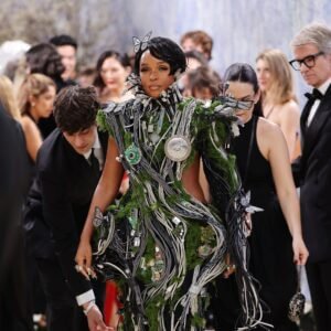 Janelle Monáe au Met Gala 2026. Elle porte une robe sculpturale complexe mêlant nature et technologie : des câbles électriques gris et noirs s'entrelacent avec de la mousse verte, des circuits imprimés et des plantes succulentes. Des papillons argentés sont posés sur l'ensemble, y compris dans ses cheveux courts et noirs.