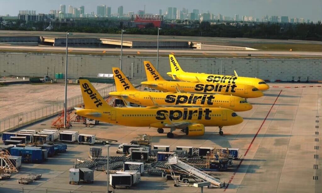 Quatre avions jaunes de la compagnie Spirit Airlines alignés sur le tarmac d'un aéroport durant la journée. En arrière-plan, on aperçoit la ligne d'horizon d'une ville avec plusieurs gratte-ciel sous un ciel nuageux. Le premier plan montre du matériel d'assistance au sol.