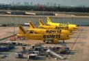 Quatre avions jaunes de la compagnie Spirit Airlines alignés sur le tarmac d'un aéroport durant la journée. En arrière-plan, on aperçoit la ligne d'horizon d'une ville avec plusieurs gratte-ciel sous un ciel nuageux. Le premier plan montre du matériel d'assistance au sol.