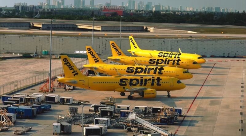 Quatre avions jaunes de la compagnie Spirit Airlines alignés sur le tarmac d'un aéroport durant la journée. En arrière-plan, on aperçoit la ligne d'horizon d'une ville avec plusieurs gratte-ciel sous un ciel nuageux. Le premier plan montre du matériel d'assistance au sol.